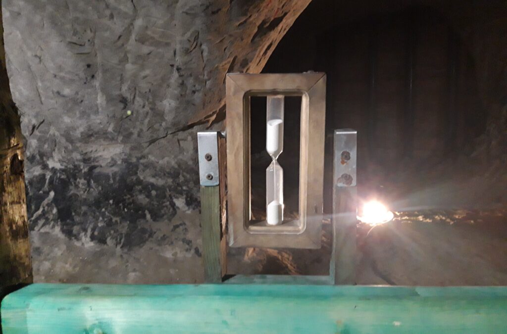 Klepsydra wypełniona piaskiem z Grot Nagórzyckich, prezentowana jako część trasy turystycznej. Symbolizuje przemijanie czasu i dawną działalność wydobywczą w Tomaszowie Mazowieckim.